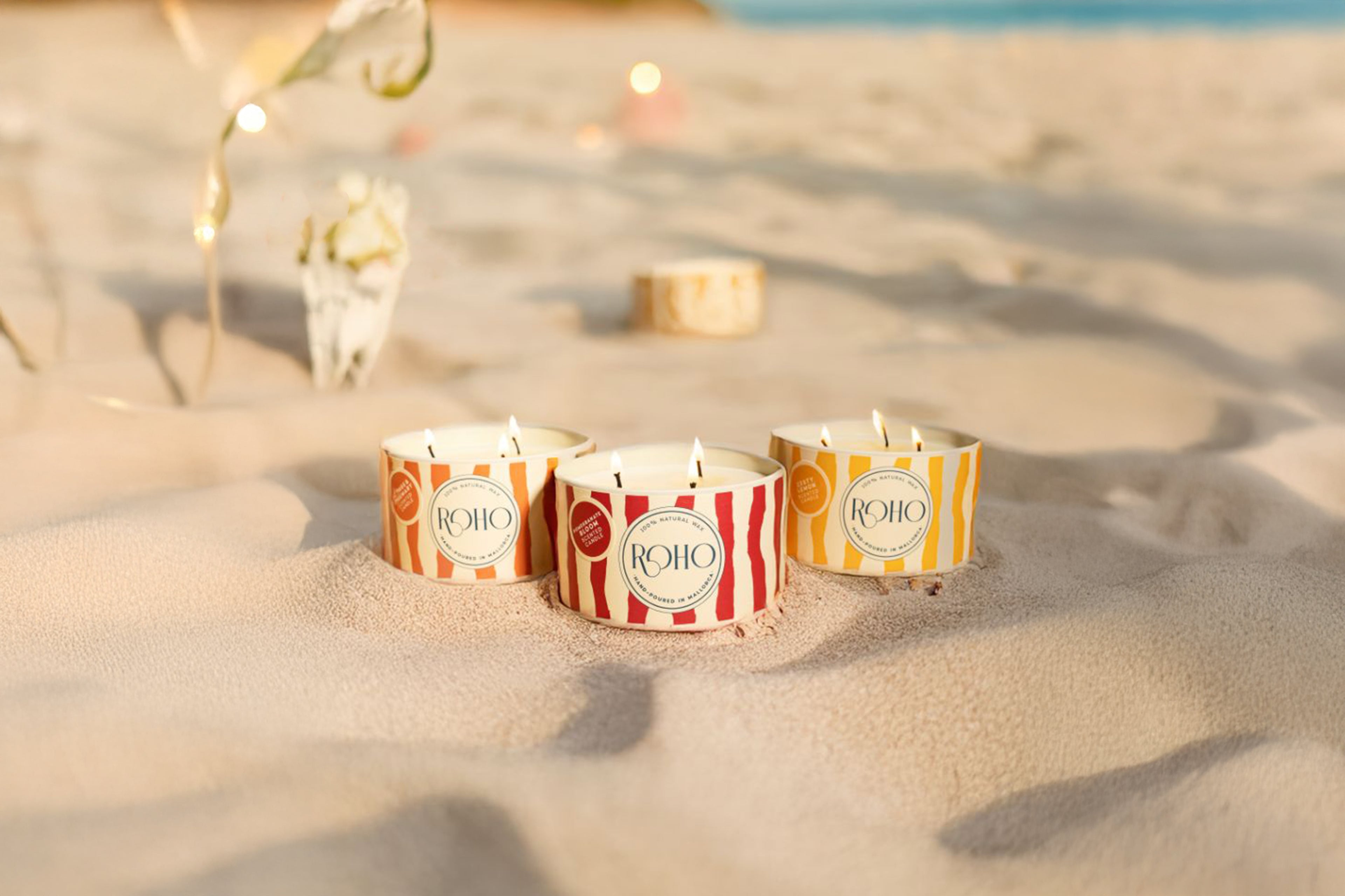 Tres velas con la marca "ROHO" sobre una superficie arenosa, rodeadas de luces de hadas.